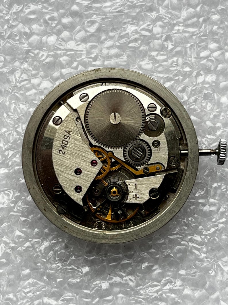 May include: Close-up of a disassembled watch movement. The silver-colored mechanism features numerous gears, springs, and other components. The watch movement has a winding stem and the number "2409A" is visible.
