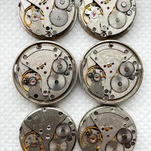 May include: Six vintage watch movements with exposed gears, springs, and jewels. The metal components are silver and gold-toned, with red jewels. The watch movements are circular and feature intricate mechanical details, showcasing the inner workings of timepieces.