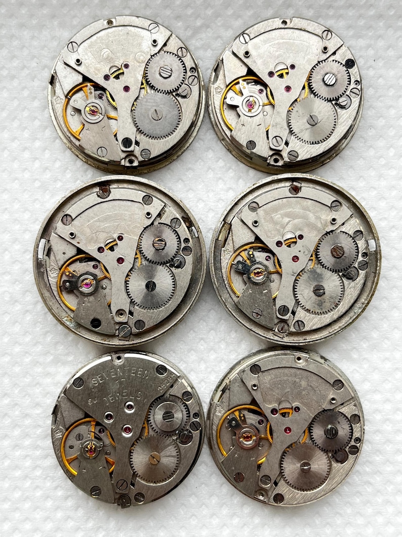 May include: Six vintage watch movements with exposed gears, springs, and jewels. The metal components are silver and gold-toned, with red jewels. The watch movements are circular and feature intricate mechanical details, showcasing the inner workings of timepieces.