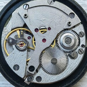 May include: Close-up of a watch mechanism with intricate gears, springs, and jewels. The metal components are silver and gold, set within a black circular frame. The detailed design showcases the inner workings of a timepiece.