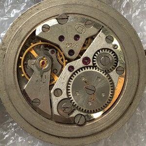 May include: Close-up view of a silver watch mechanism with intricate gears and a winding stem. The mechanism is exposed and visible through a clear glass face.