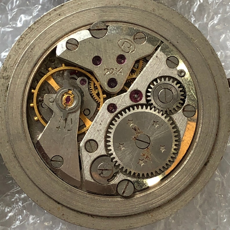 May include: Close-up view of a silver watch mechanism with intricate gears and a winding stem. The mechanism is exposed and visible through a clear glass face.