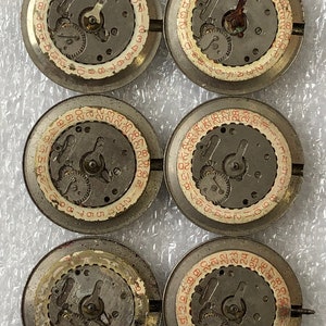 May include: Five round, silver-coloured watch parts with red numbers and black gears. The parts are arranged in a circle on a white surface.