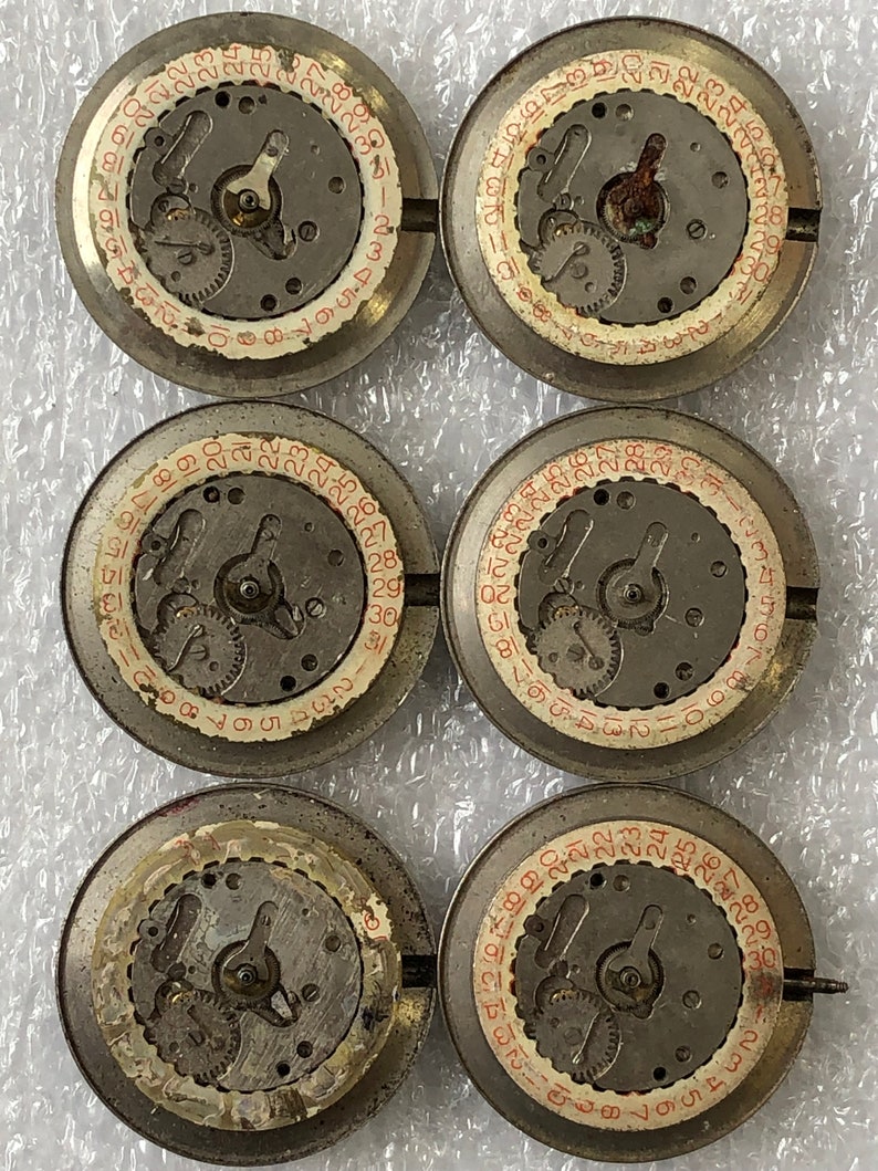 May include: Five round, silver-coloured watch parts with red numbers and black gears. The parts are arranged in a circle on a white surface.