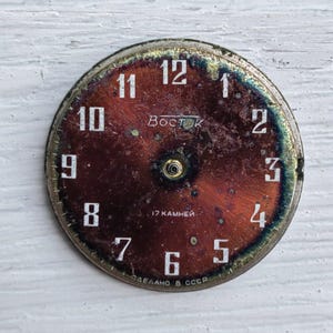 May include: Vintage watch face with a dark red and bronze color scheme. White numerals mark the hours, with the brand name "Восток" and text "17 камней" and "СДЕЛАНО В СССР" visible. The face has a circular shape with a metallic rim.