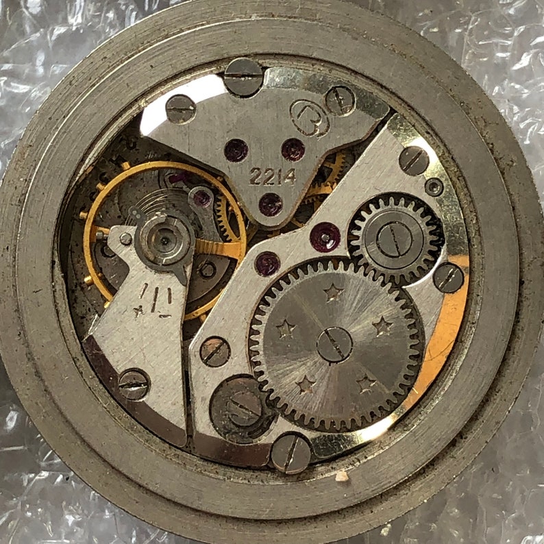 May include: Close-up view of a silver watch mechanism with gold gears and a black dial. The watch face has the number 2214 printed on it.