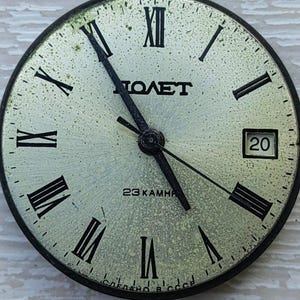May include: Vintage watch face with black Roman numerals and hands on a speckled, light-colored background. The watch face has the word "POLET" in the center, with the date 20 visible. The text "23 KAMHЯ" and "СДЕЛАНО В СССР" are also present.