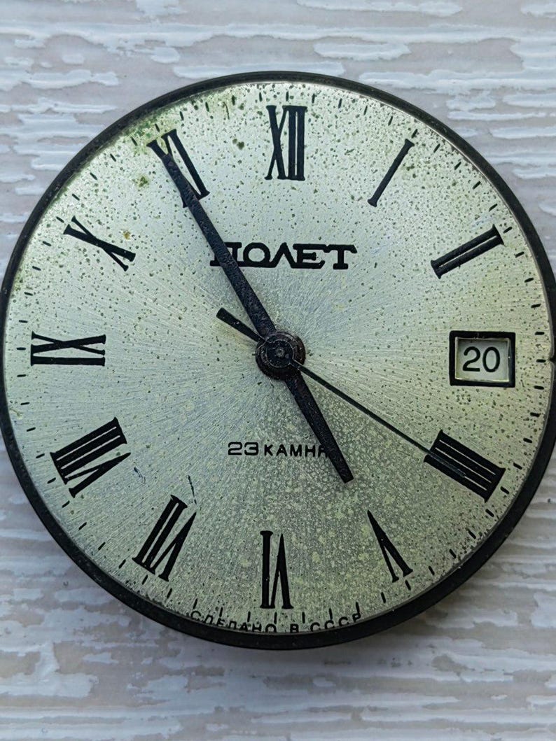 May include: Vintage watch face with black Roman numerals and hands on a speckled, light-colored background. The watch face has the word "POLET" in the center, with the date 20 visible. The text "23 KAMHЯ" and "СДЕЛАНО В СССР" are also present.