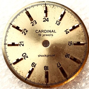 Puede incluir: Una esfera de reloj de color dorado con marcas negras y el texto "CARDINAL 19 jewels" y "shockproof". Los números del 13 al 24 se muestran alrededor del borde de la esfera.