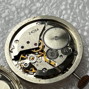 May include: Close-up of a watch movement with visible gears, springs, and the text "2409A". The intricate mechanical components are housed in a silver-colored casing. The watch's crown is visible on the right side.