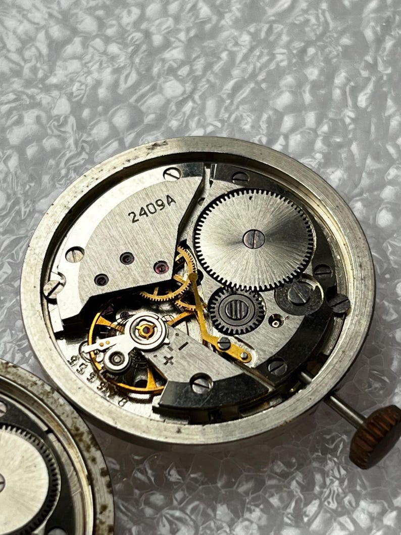 May include: Close-up of a watch movement with visible gears, springs, and the text "2409A". The intricate mechanical components are housed in a silver-colored casing. The watch's crown is visible on the right side.