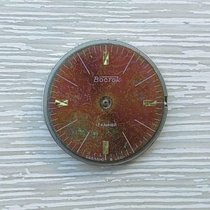 May include: A vintage watch face with a reddish-brown dial and gold-colored hour markers. The word "Vostok" is printed in the center. The watch face has a silver-colored metal rim and is set against a light-colored, textured background.