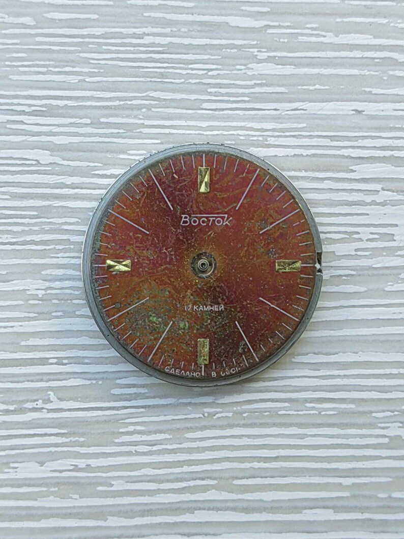 May include: A vintage watch face with a reddish-brown dial and gold-colored hour markers. The word "Vostok" is printed in the center. The watch face has a silver-colored metal rim and is set against a light-colored, textured background.