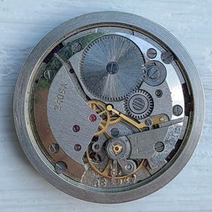 May include: Close-up of a vintage watch movement with intricate gears, screws, and a balance wheel. The metal components are silver and gold, with the text "240SA" visible. The watch mechanism is housed in a circular frame.