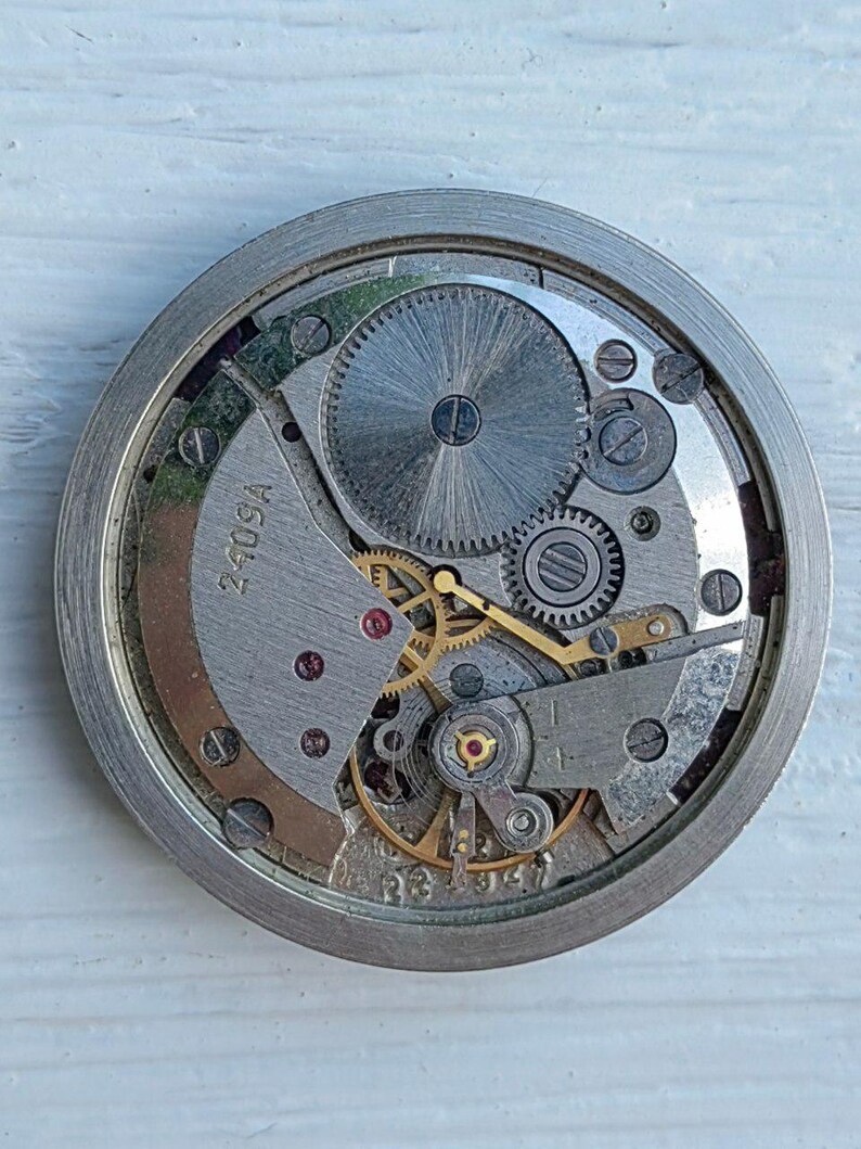 May include: Close-up of a vintage watch movement with intricate gears, screws, and a balance wheel. The metal components are silver and gold, with the text "240SA" visible. The watch mechanism is housed in a circular frame.