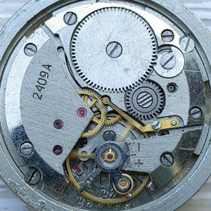 K&ouml;nnte beinhalten: Nahaufnahme eines Uhrwerks mit komplexen Zahnr&auml;dern, Schrauben und einer Unruh. Die Metallkomponenten sind silber- und goldfarben, mit dem Text "2409A" auf einer Platte.