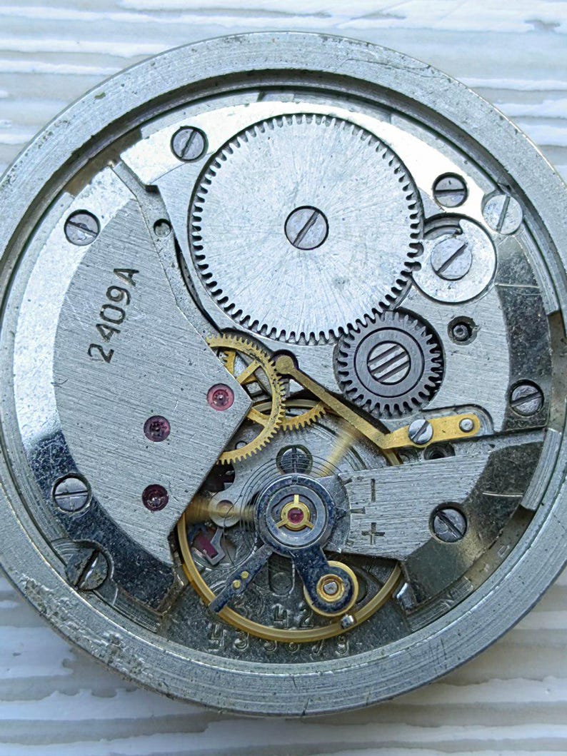 K&ouml;nnte beinhalten: Nahaufnahme eines Uhrwerks mit komplexen Zahnr&auml;dern, Schrauben und einer Unruh. Die Metallkomponenten sind silber- und goldfarben, mit dem Text "2409A" auf einer Platte.