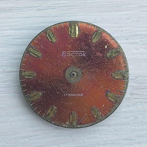 K&ouml;nnte beinhalten: Ein Vintage-Zifferblatt in rotbrauner Farbe mit goldfarbenen Stundenmarkierungen und Zeigern. Das Wort "Восток" ist in Wei&szlig; gedruckt, zusammen mit "17 камней" und "A CCCP".