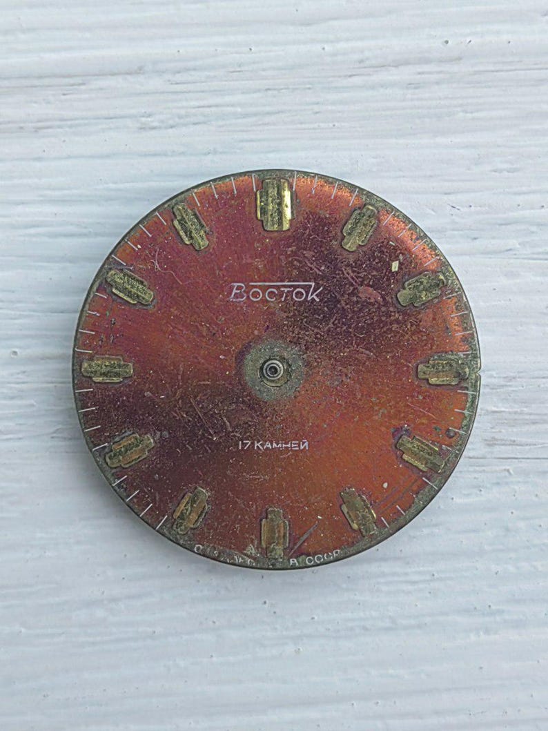 K&ouml;nnte beinhalten: Ein Vintage-Zifferblatt in rotbrauner Farbe mit goldfarbenen Stundenmarkierungen und Zeigern. Das Wort "Восток" ist in Wei&szlig; gedruckt, zusammen mit "17 камней" und "A CCCP".