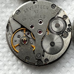 May include: Close-up of a vintage watch movement. The silver-colored mechanism features the text "SEVENTEEN 17 JEWELS". Visible are gears, screws, and ruby jewels. The intricate design showcases the inner workings of a classic timepiece.