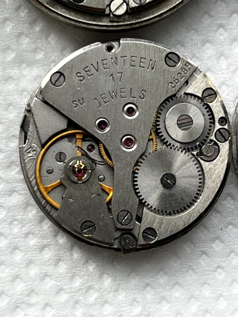 May include: Close-up of a vintage watch movement. The silver-colored mechanism features the text "SEVENTEEN 17 JEWELS". Visible are gears, screws, and ruby jewels. The intricate design showcases the inner workings of a classic timepiece.