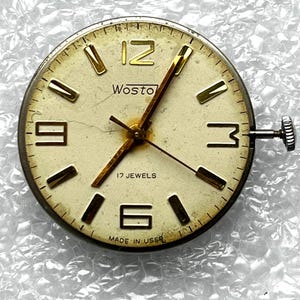 May include: Vintage Wostok watch face with a cream-colored dial, gold-tone hands and markers, and the text "17 JEWELS" and "MADE IN USSR". The watch face has a silver-tone bezel and a winding crown. The watch face is round.