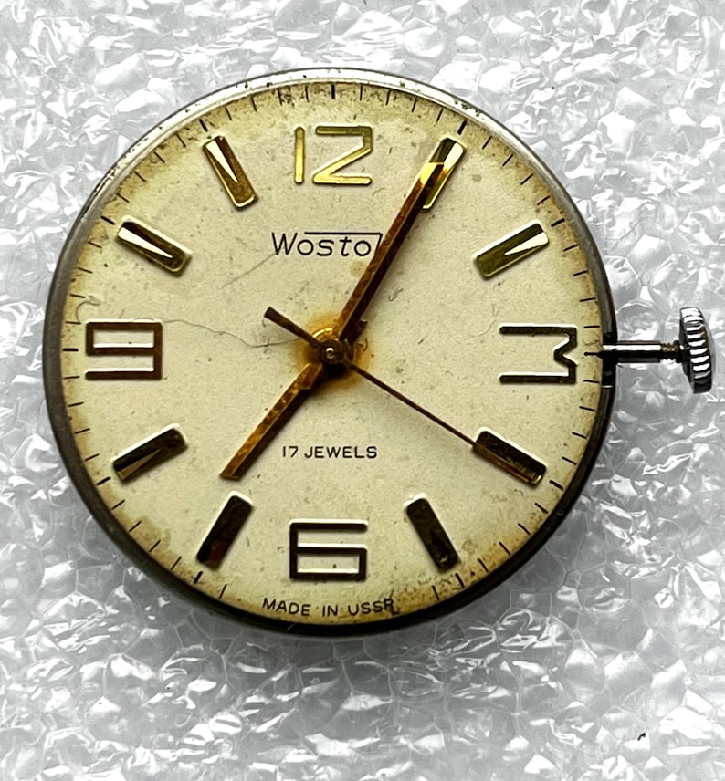 May include: Vintage Wostok watch face with a cream-colored dial, gold-tone hands and markers, and the text "17 JEWELS" and "MADE IN USSR". The watch face has a silver-tone bezel and a winding crown. The watch face is round.