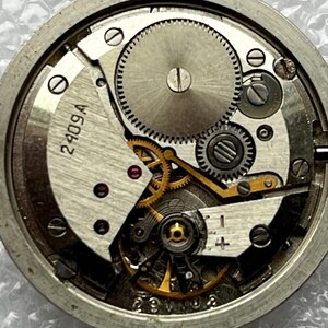 May include: Close-up of a watch movement with intricate gears and components. The metal parts are silver and gold, with the text "2409A" visible. The watch mechanism is a complex arrangement of small parts.
