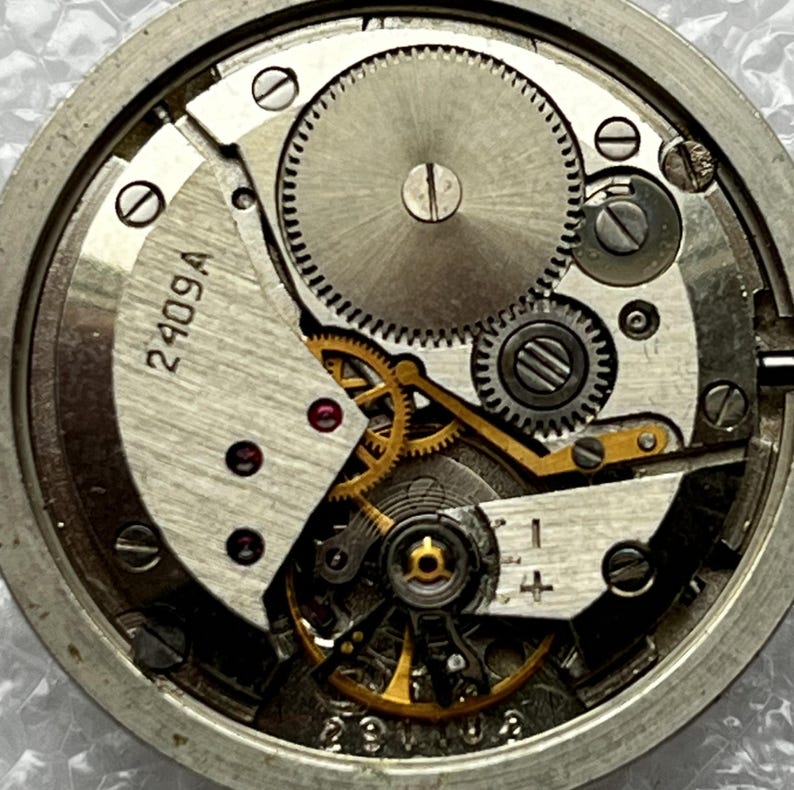 May include: Close-up of a watch movement with intricate gears and components. The metal parts are silver and gold, with the text "2409A" visible. The watch mechanism is a complex arrangement of small parts.