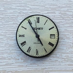 May include: A vintage watch face with a light green background, black Roman numerals, and black hands. The watch face has the word "ПОЛЕТ" printed in black, along with the number "20" and the text "23 KAMHЯ".