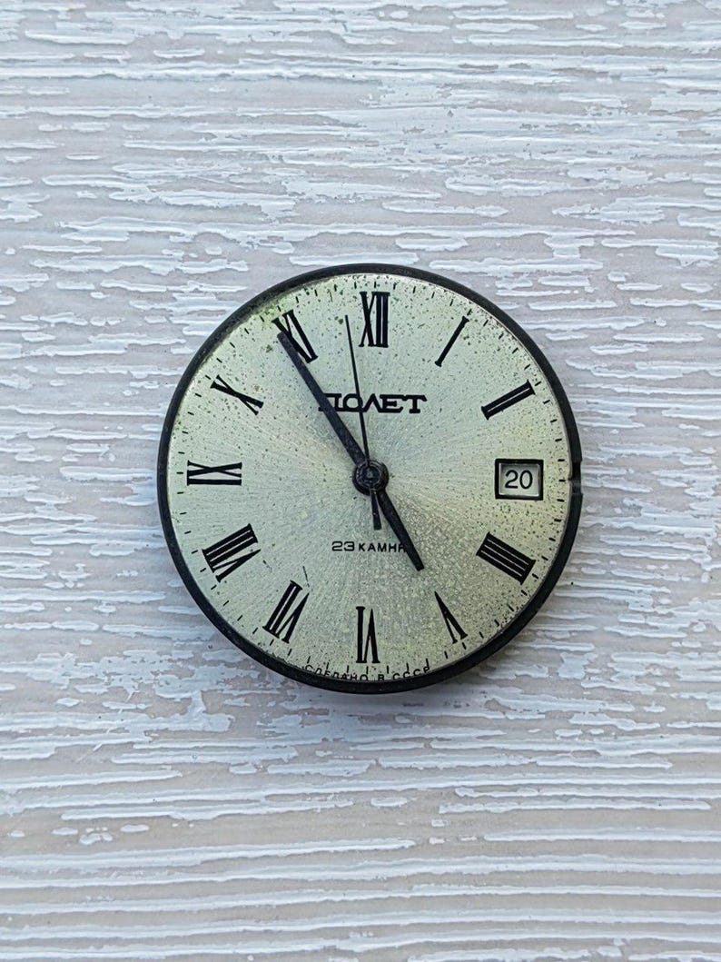 May include: A vintage watch face with a light green background, black Roman numerals, and black hands. The watch face has the word "ПОЛЕТ" printed in black, along with the number "20" and the text "23 KAMHЯ".