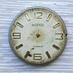 K&ouml;nnte beinhalten: Vintage Wostok Zifferblatt mit cremefarbenem Zifferblatt und goldfarbenen Ziffern. Das Zifferblatt tr&auml;gt die Aufschrift "Wostok" und "17 Jewels". Die Worte "Made in USA" sind ebenfalls sichtbar. Das Zifferblatt ist rund und hat einen silberfarbenen Metallrahmen.