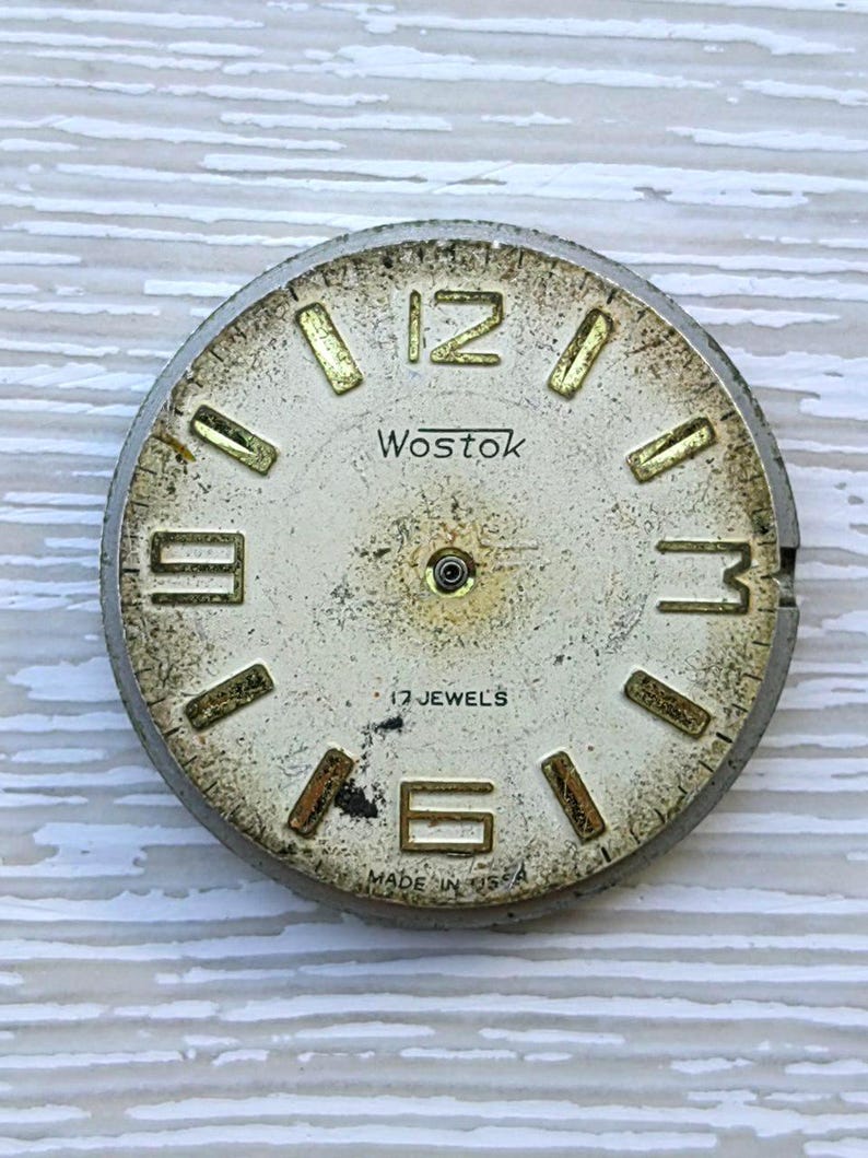 K&ouml;nnte beinhalten: Vintage Wostok Zifferblatt mit cremefarbenem Zifferblatt und goldfarbenen Ziffern. Das Zifferblatt tr&auml;gt die Aufschrift "Wostok" und "17 Jewels". Die Worte "Made in USA" sind ebenfalls sichtbar. Das Zifferblatt ist rund und hat einen silberfarbenen Metallrahmen.