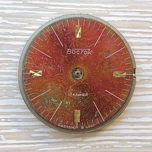 May include: Vintage watch face with a red and gold dial. The face features the brand name "Vostok" and the text "17 камней". The dial has gold-colored rectangular markers and white lines.