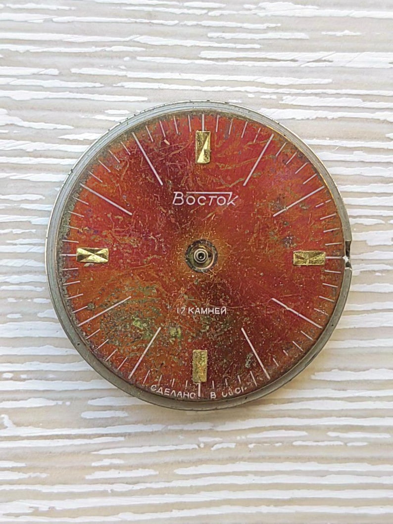 May include: Vintage watch face with a red and gold dial. The face features the brand name "Vostok" and the text "17 камней". The dial has gold-colored rectangular markers and white lines.