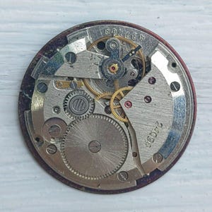 K&ouml;nnte beinhalten: Nahaufnahme eines Vintage-Uhrwerks, das die komplizierten Zahnr&auml;der, Federn und andere mechanische Komponenten zeigt. Der Uhrenmechanismus befindet sich vor einem kreisf&ouml;rmigen, dunkelroten Hintergrund, auf dem die Zahlen "437268" und "2409A" zu sehen sind.