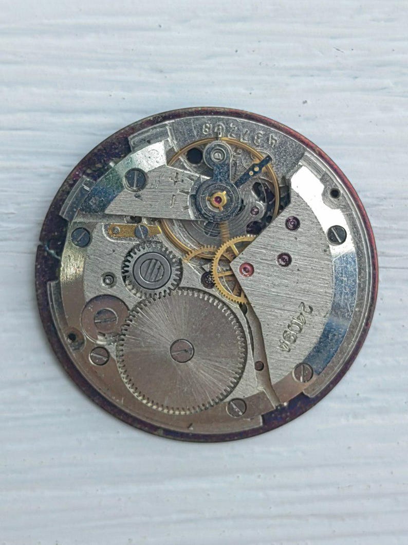 K&ouml;nnte beinhalten: Nahaufnahme eines Vintage-Uhrwerks, das die komplizierten Zahnr&auml;der, Federn und andere mechanische Komponenten zeigt. Der Uhrenmechanismus befindet sich vor einem kreisf&ouml;rmigen, dunkelroten Hintergrund, auf dem die Zahlen "437268" und "2409A" zu sehen sind.