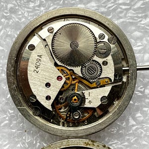 May include: Close-up of a disassembled watch movement, showcasing intricate gears, springs, and the text "2409A" on a white component. The metal components are silver and gold, with a circular design.
