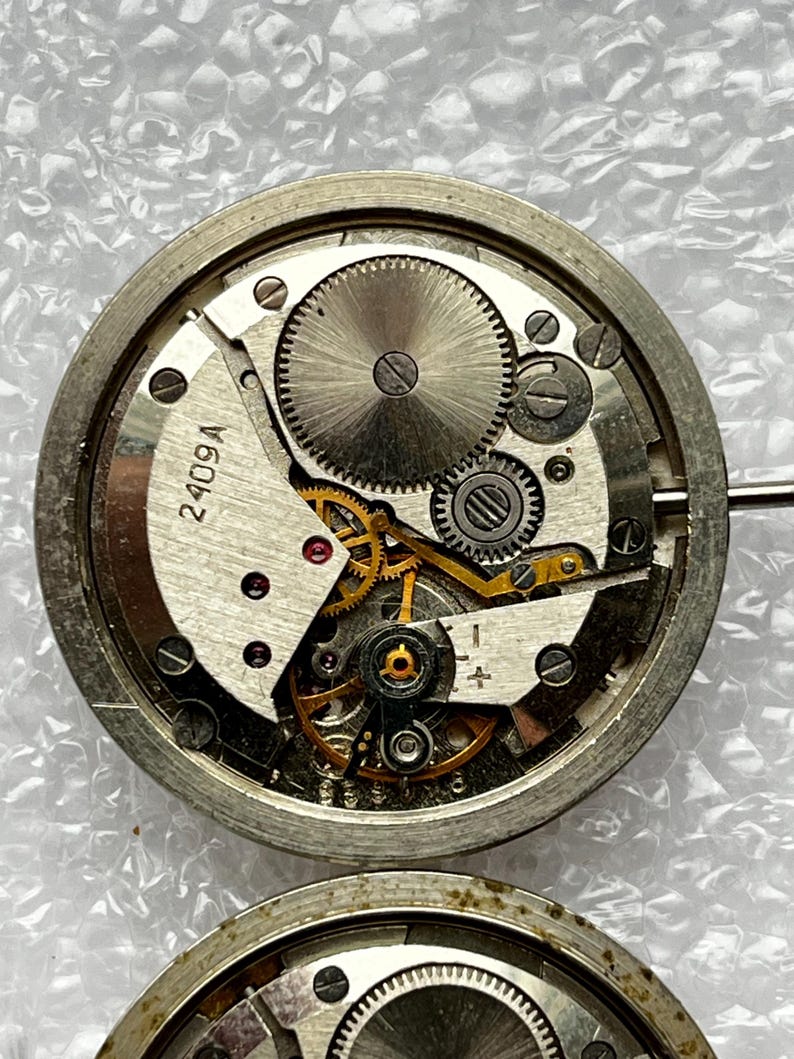 May include: Close-up of a disassembled watch movement, showcasing intricate gears, springs, and the text "2409A" on a white component. The metal components are silver and gold, with a circular design.