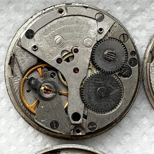 May include: Close-up of a disassembled watch movement. The intricate mechanism features silver-colored metal components, including gears, springs, and screws. A gold-colored balance wheel and ruby jewels are also visible. The watch movement is circular.