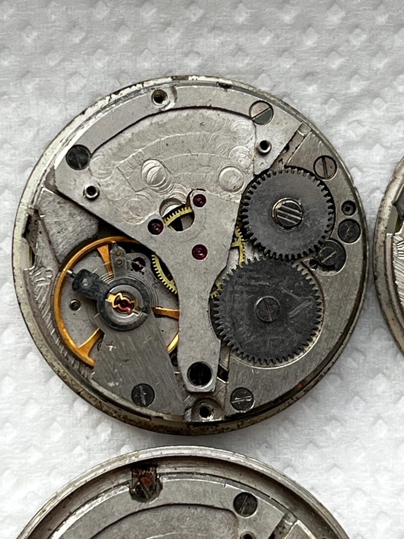 May include: Close-up of a disassembled watch movement. The intricate mechanism features silver-colored metal components, including gears, springs, and screws. A gold-colored balance wheel and ruby jewels are also visible. The watch movement is circular.
