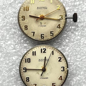 May include: Two vintage watch faces with cream-colored dials. The faces feature gold-toned numerals, hands, and the word "Восток". The text "17 KAMHEЙ" and "СДЕЛАНО В СССР" are also visible. One face has a stem and crown.