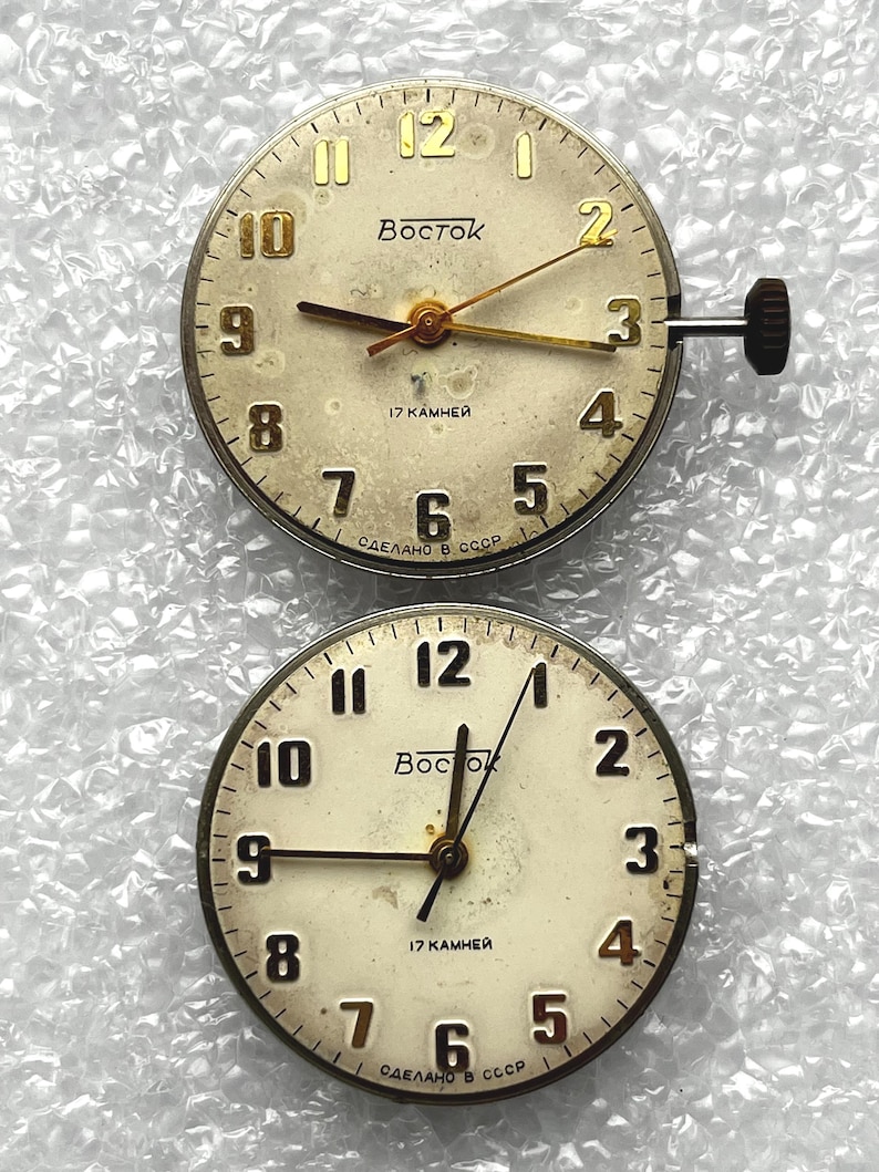May include: Two vintage watch faces with cream-colored dials. The faces feature gold-toned numerals, hands, and the word "Восток". The text "17 KAMHEЙ" and "СДЕЛАНО В СССР" are also visible. One face has a stem and crown.