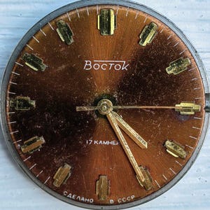 May include: Vintage Vostok watch face with a copper-colored dial. The face features gold-toned hour markers and hands. The word "Vostok" is printed in white. The text "17 KAMHEЙ" and "СДЕЛАНО В СССР" are also visible.