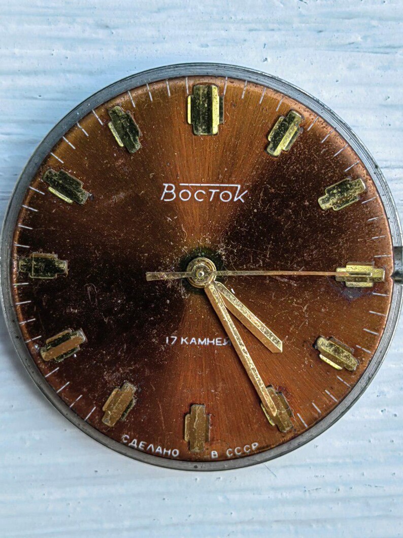 May include: Vintage Vostok watch face with a copper-colored dial. The face features gold-toned hour markers and hands. The word "Vostok" is printed in white. The text "17 KAMHEЙ" and "СДЕЛАНО В СССР" are also visible.