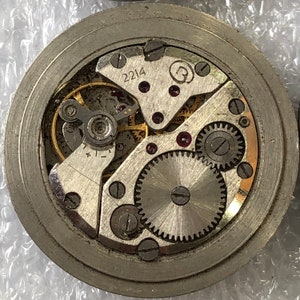 May include: Close-up view of a silver watch mechanism with exposed gears and a circular dial labelled "2214" and "R".