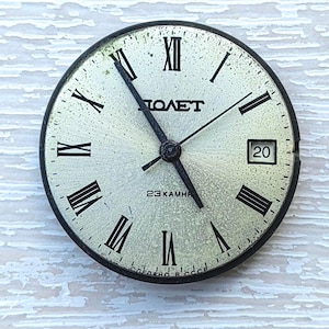 May include: A vintage watch face with a silver-colored dial, black Roman numerals, and black hands. The watch face has the word "ПОЛЕТ" and "23 KAMHЯ" printed on it. The date is displayed at the 3 o'clock position.