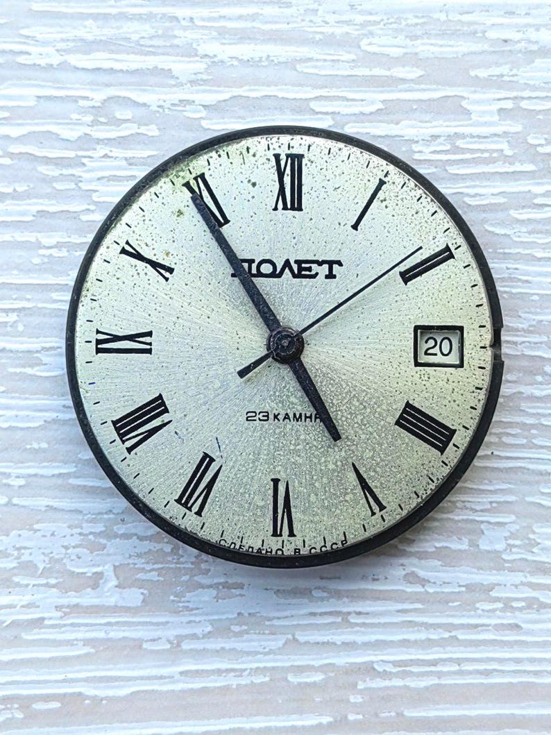May include: A vintage watch face with a silver-colored dial, black Roman numerals, and black hands. The watch face has the word "ПОЛЕТ" and "23 KAMHЯ" printed on it. The date is displayed at the 3 o'clock position.