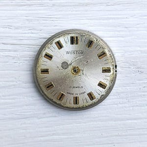 May include: A vintage watch face with a silver metal frame. The face is a light gold colour with black and gold rectangular hour markers. The text "Wostok", "17 Jewels", and "Made in USSR" are printed on the face.