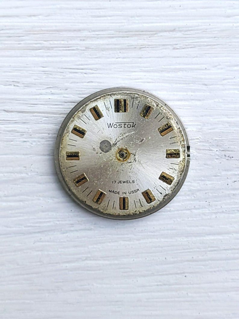 May include: A vintage watch face with a silver metal frame. The face is a light gold colour with black and gold rectangular hour markers. The text "Wostok", "17 Jewels", and "Made in USSR" are printed on the face.